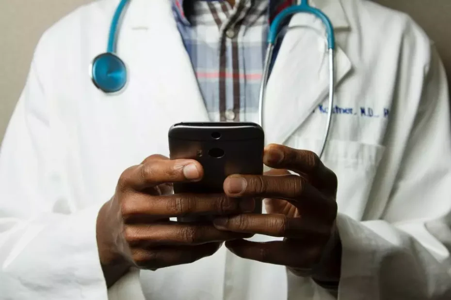 A doctor uses a cellphone. They have a stethoscope hanging around their neck and are wearing a lab coat.