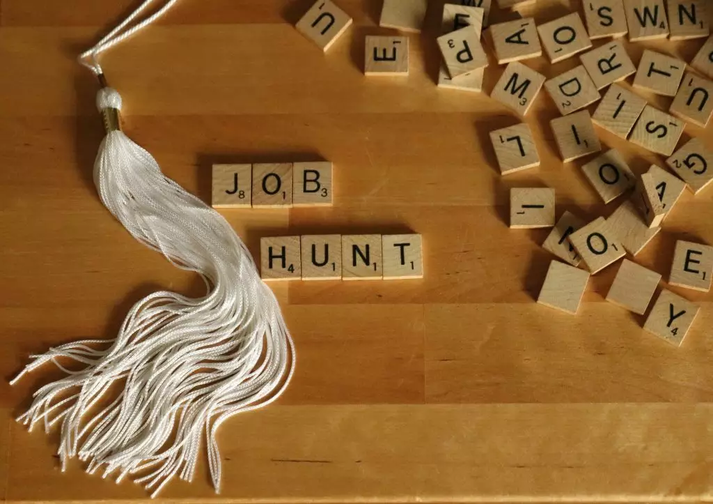 JOB HUNT is spelled out in simple wooden tiles beside a white graduation cap tassle. A pile of jumbled wooden letters decorate the right side against a wooden butcherblock background.