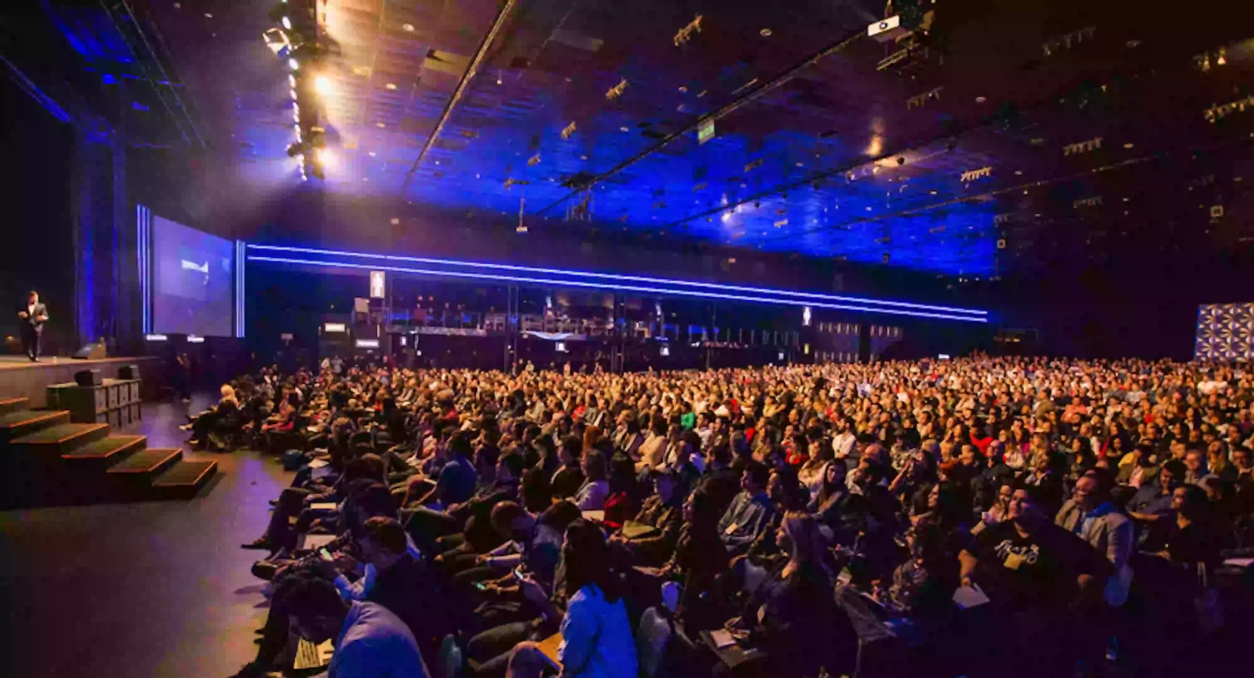 A crowd of people at a large conference venue are bathed in blue light.